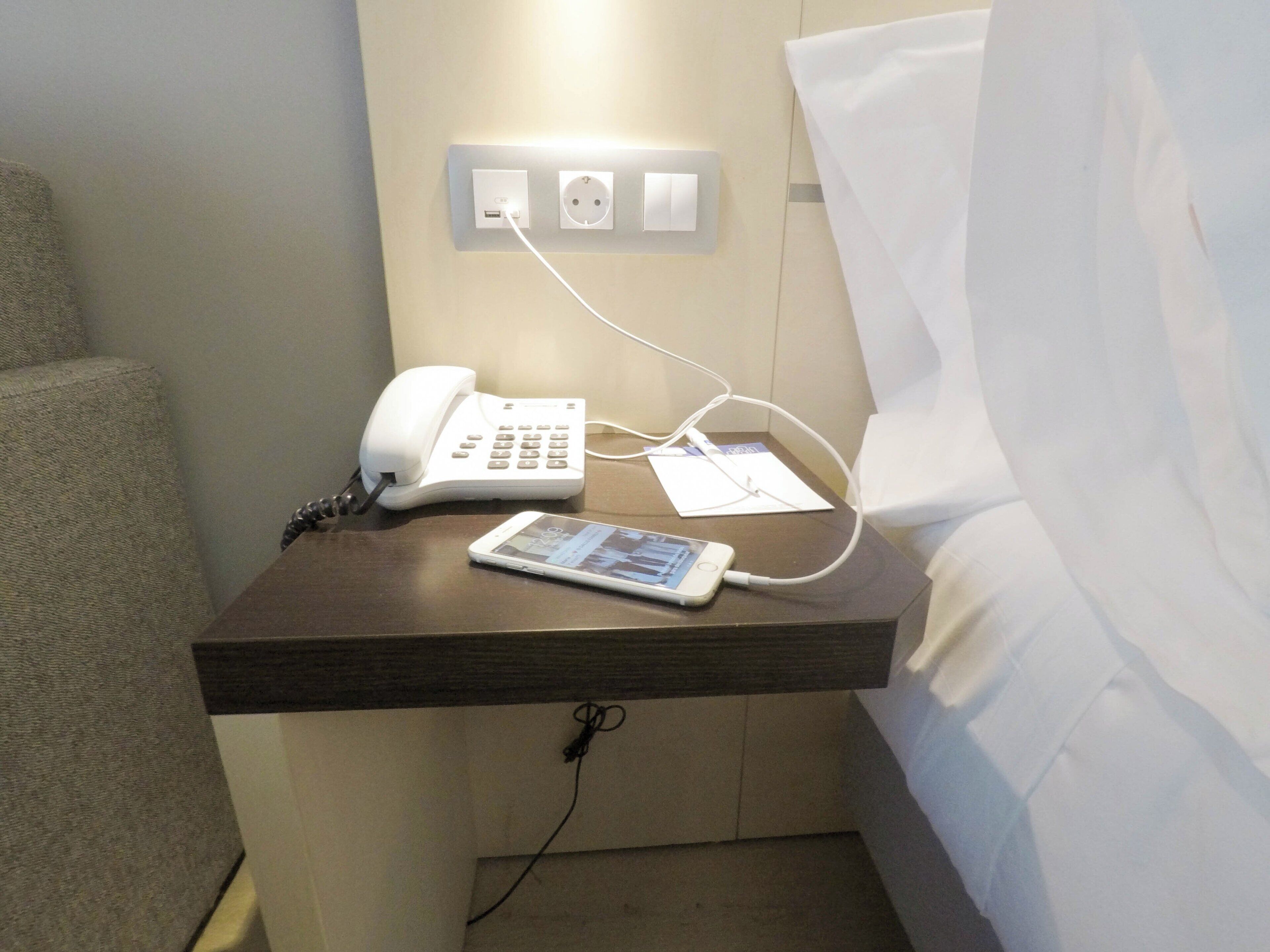 Una camera d'albergo con un comodino su cui sono posizionati un telefono e un tablet, una presa di corrente e una presa a muro.