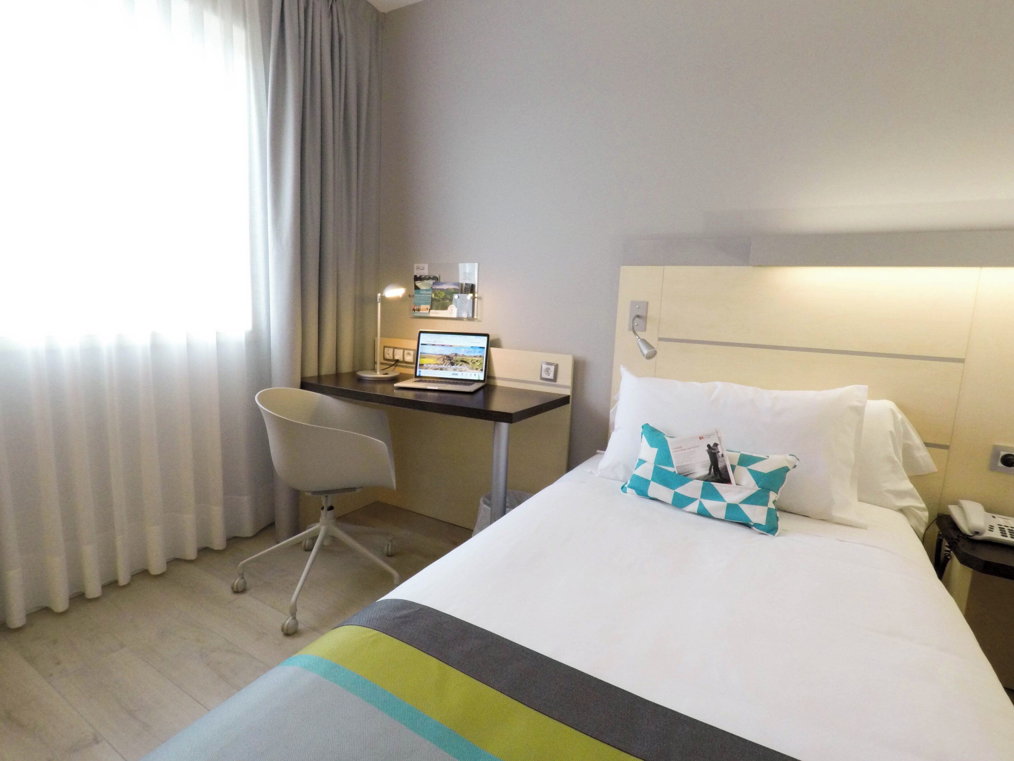 A hotel room with a bed, a desk with a laptop, a chair, and a nightstand with a phone.