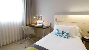 A hotel room with a bed, a desk with a laptop, a chair, and a nightstand with a phone.