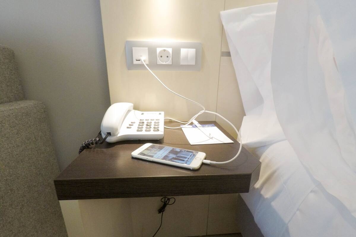 A hotel room with a bedside table holding a white phone and a tablet connected to a power outlet.