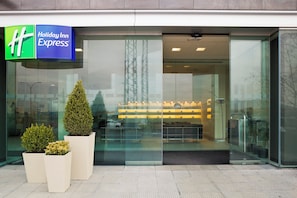 Entrada de un hotel Holiday Inn Express con fachada de vidrio, plantas en macetas y un interior visible con asientos.