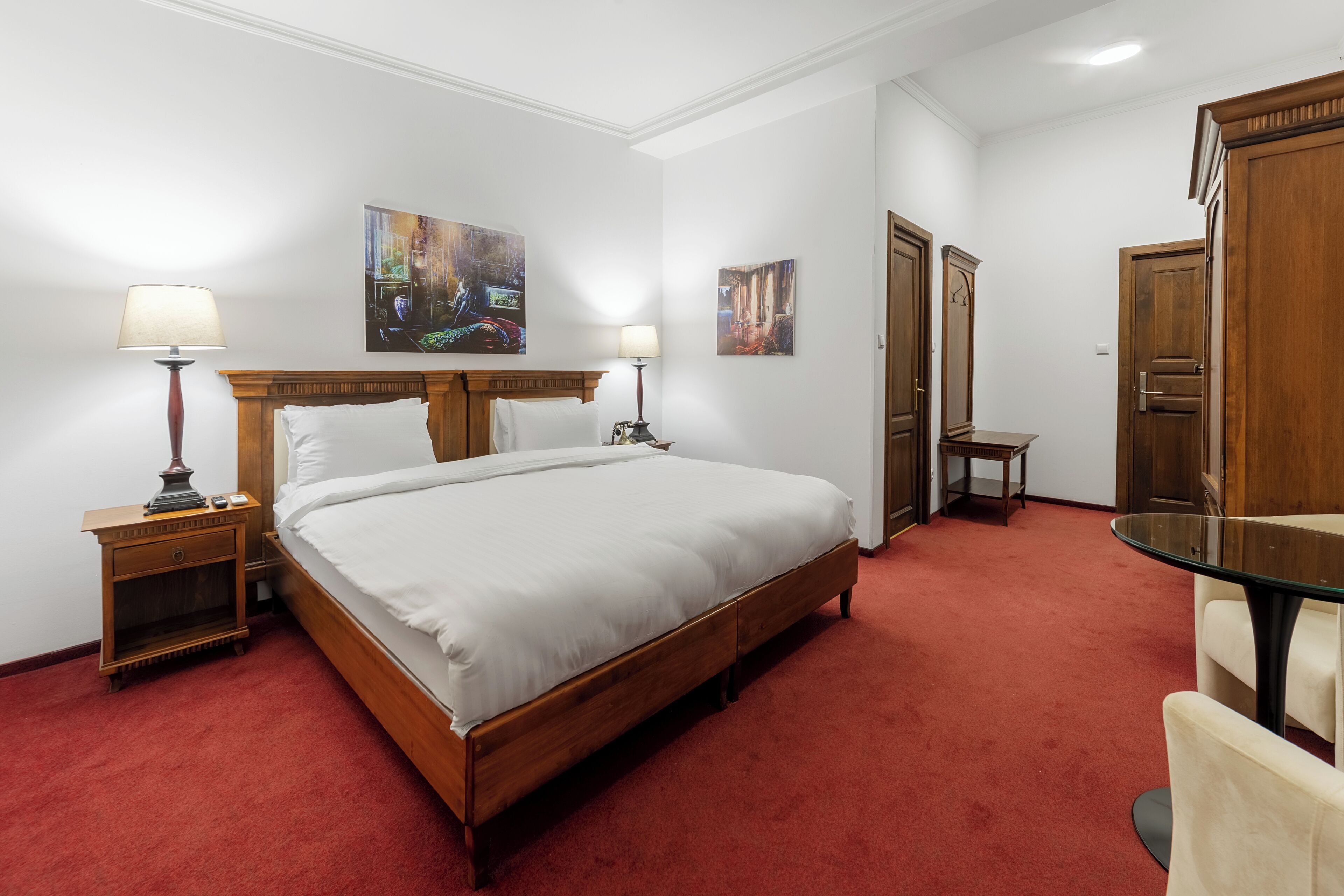 A hotel room with a large bed, two bedside tables with lamps, a small table, and a glass table with chairs.