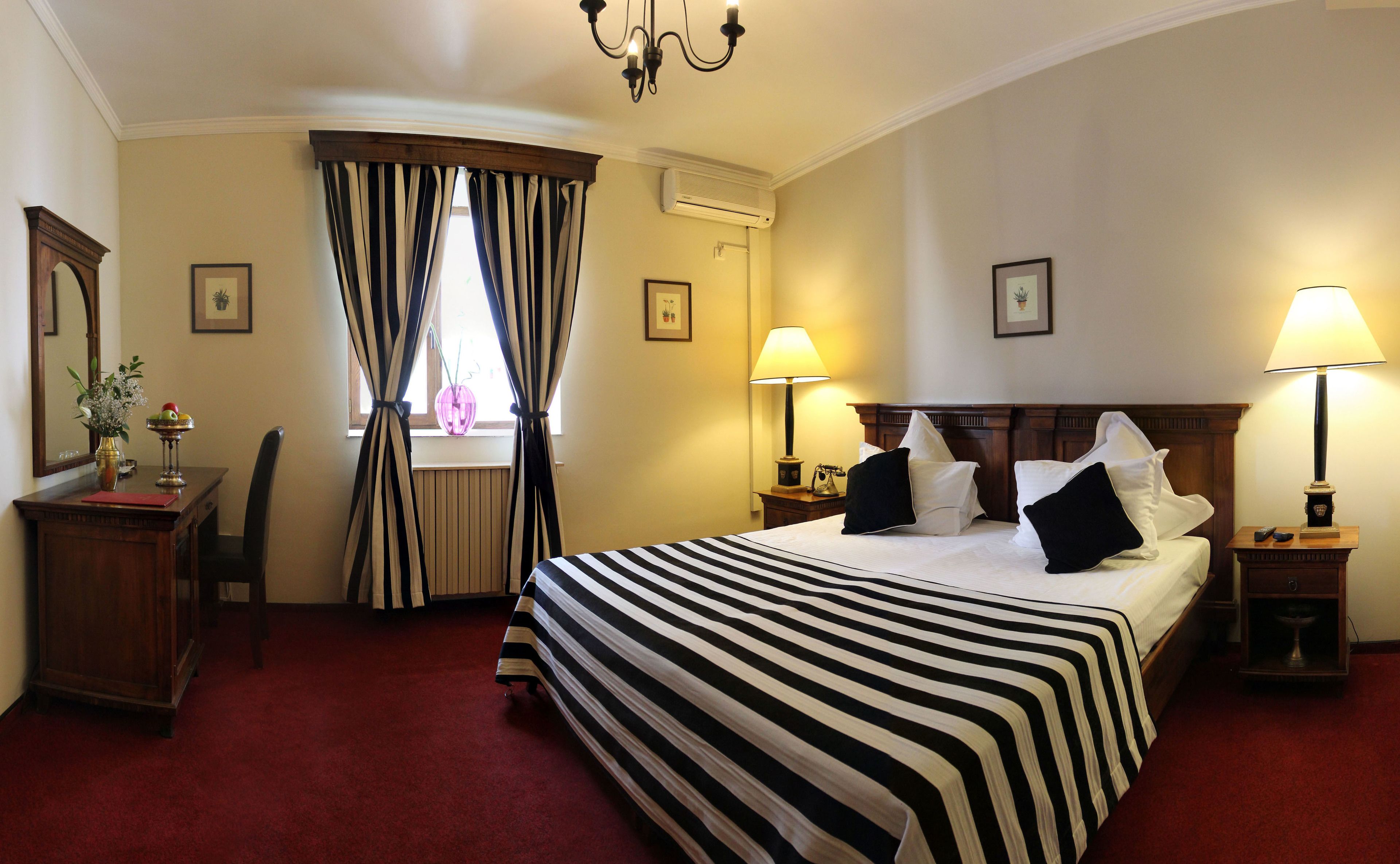 A hotel room with a large bed, two bedside lamps, a desk with a chair, a window with curtains, and a chandelier.