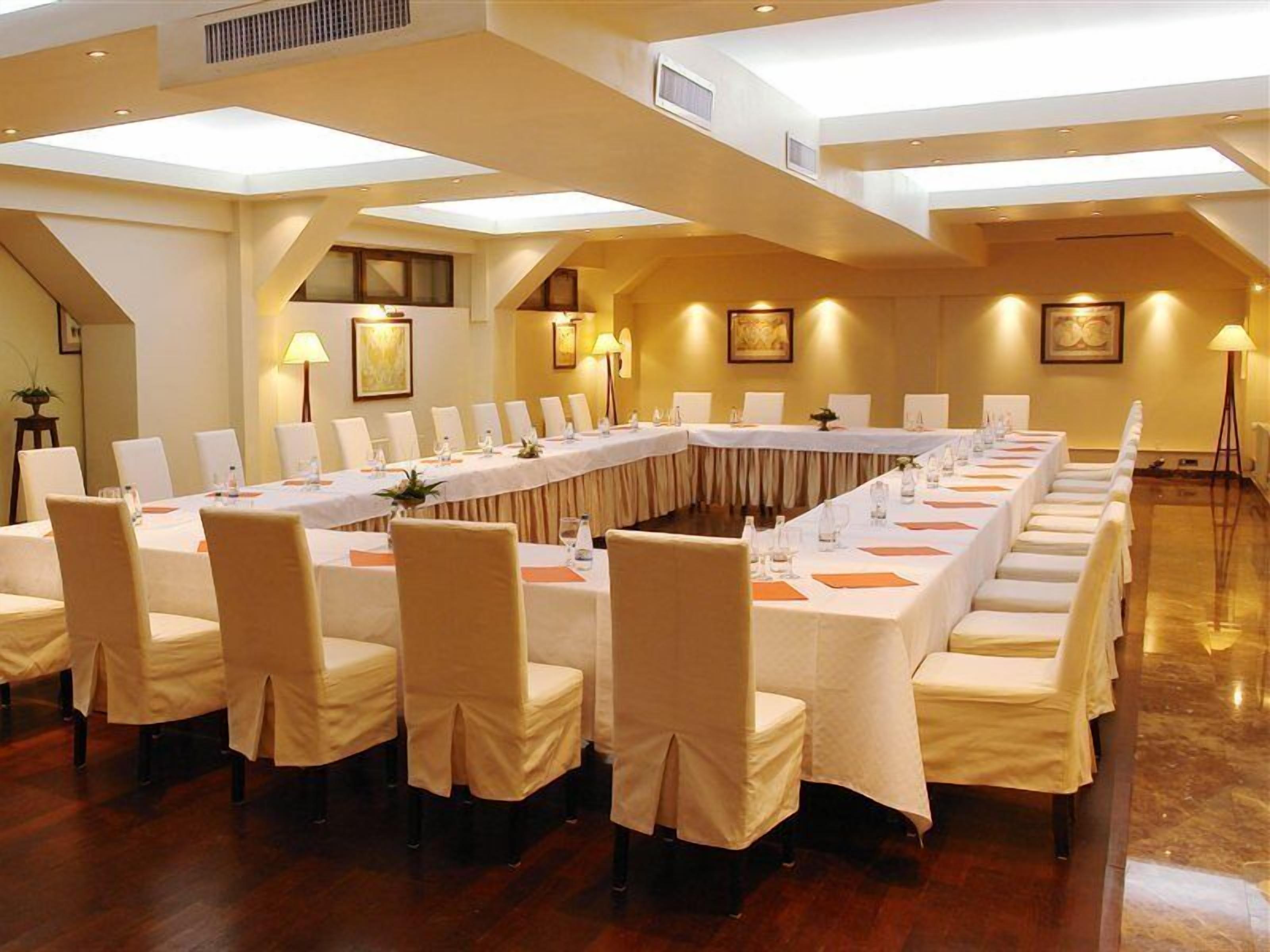 Een congreszaal met een lange tafel, stoelen en een hoge plafond met dakramen.
