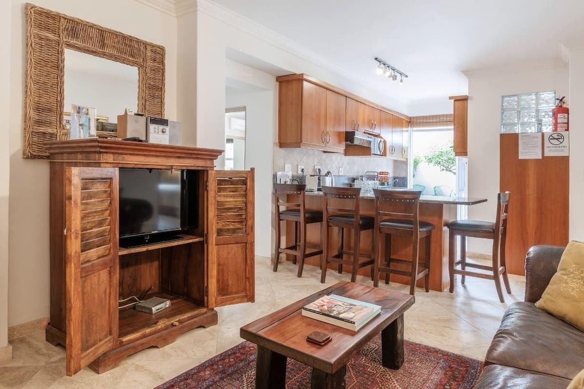 A living room with a wooden entertainment unit, a television, and a coffee table. There is a kitchen in the background with wooden cabinets and a dining area.