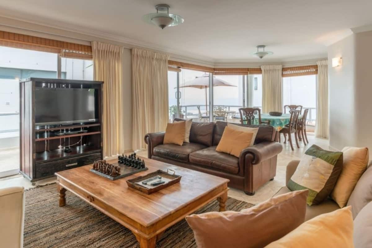 A living room with a wooden coffee table, leather sofa, and a TV. There is a balcony with a table and chairs.