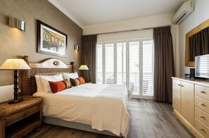 A hotel room with a large bed, a wooden nightstand, a television, and a window with curtains. - Bantry Beach Luxury Suites (Cape Town)