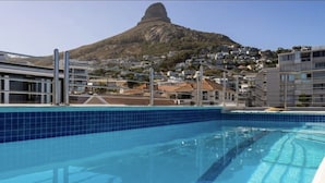 En swimmingpool med krystalklart, blåt vand, omgivet af moderne bygninger og et markant bjerg i baggrunden.