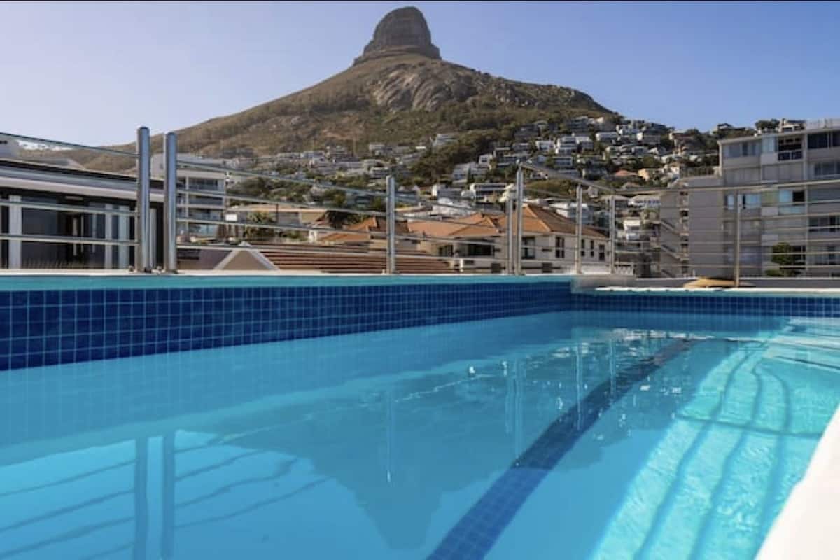 A swimming pool with clear blue water, surrounded by modern buildings and a prominent mountain in the background.