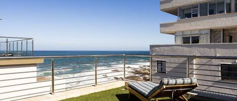 A balcony with a view of the ocean, a lounge chair, and a building with multiple balconies.