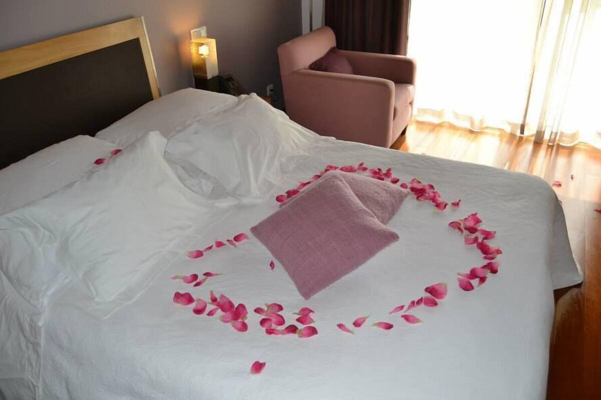 Een netjes opgemaakt bed met witte linnens, roze rozenblaadjes gerangschikt in een hartvorm, en een roze handdoek op het bed.