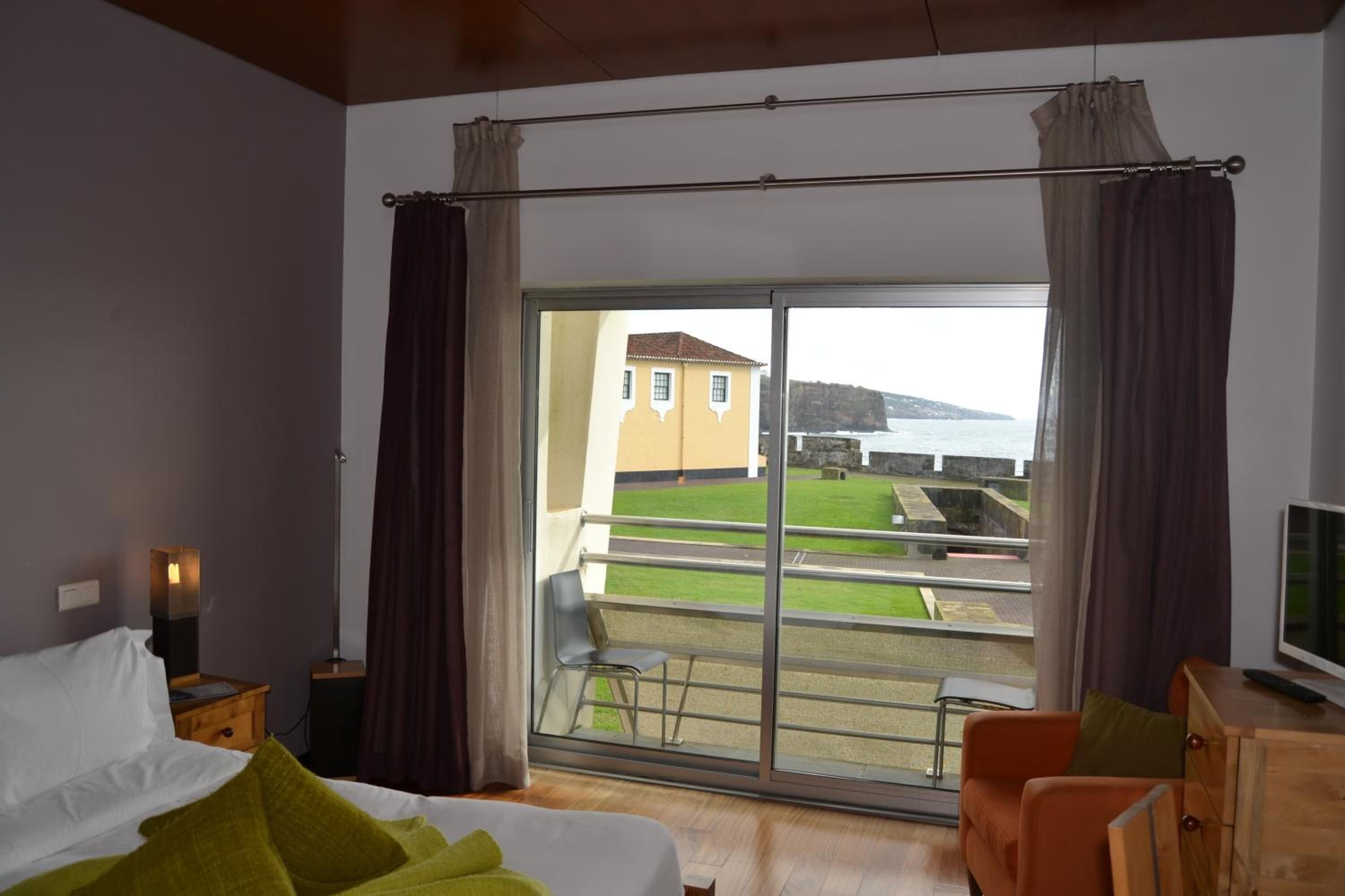 A hotel room with a bed, a TV, a balcony with a view of the sea and a building.