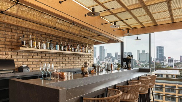 Un bar moderno con vistas a la ciudad, que presenta una pared de ladrillo, una barra larga y sillas de mimbre.