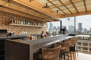 Un bar moderno con vistas a la ciudad, que presenta una pared de ladrillo, una barra larga y sillas de mimbre.