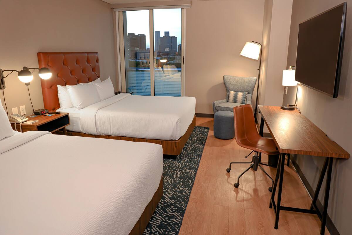 Habitación de hotel con dos camas, un escritorio, una silla y vistas de la ciudad a través de amplias ventanas.