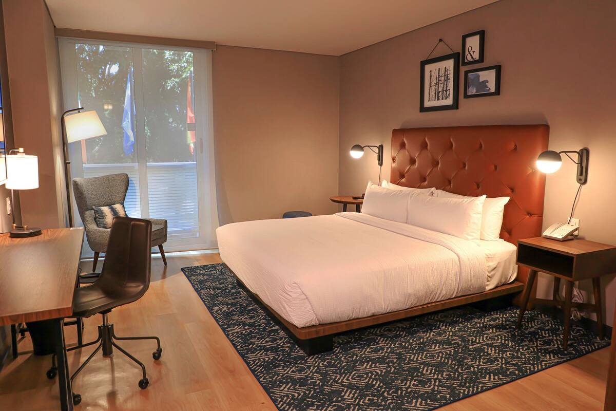 Una habitación de hotel moderna con una cama grande, un escritorio con silla, una mesita pequeña y una mesita de noche con lámpara.