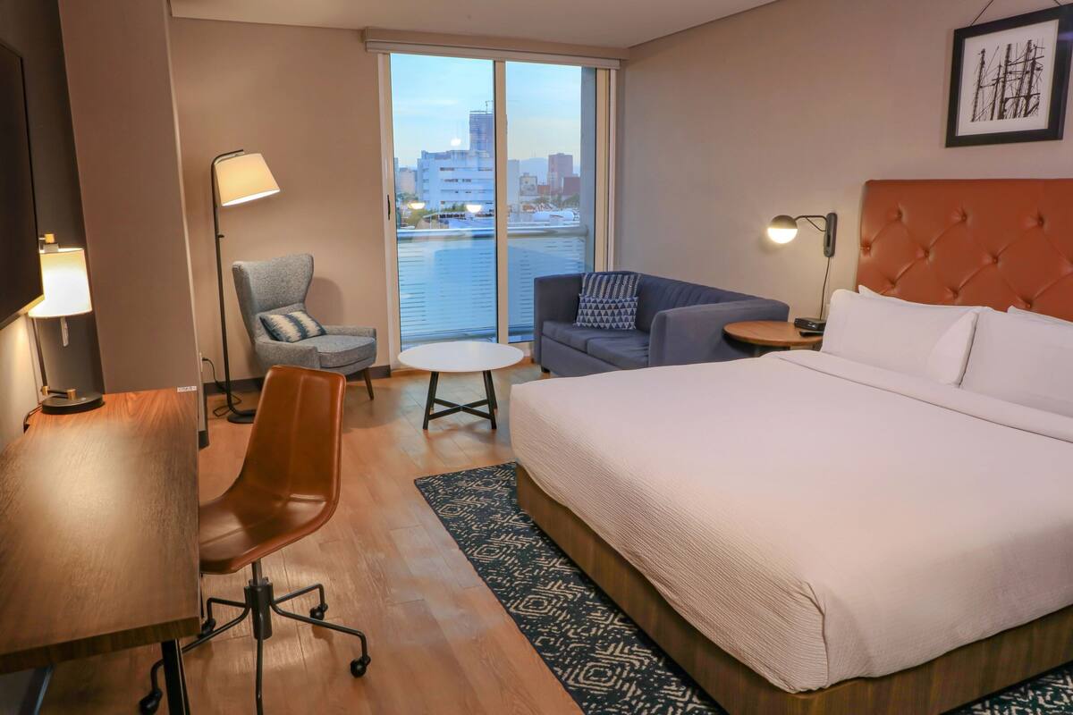 Habitación de hotel con una cama grande, un escritorio, una silla, un sofá y vistas a la ciudad desde la ventana.