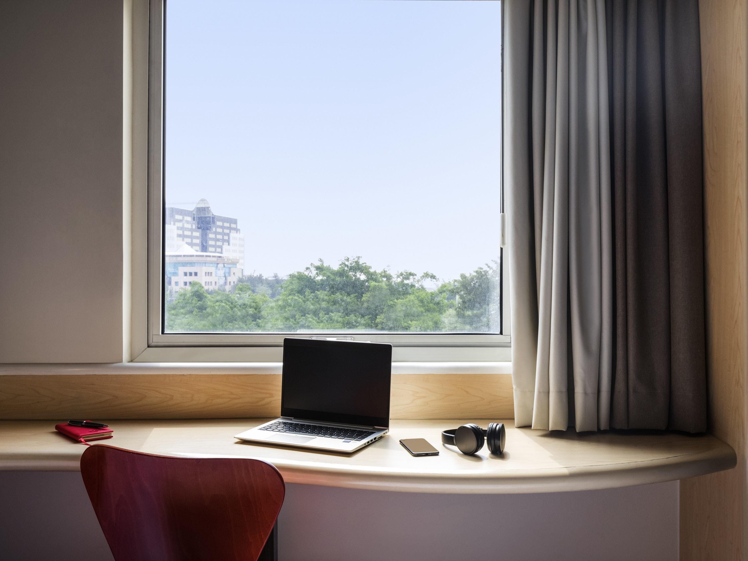 A laptop on a desk by a window with a view of buildings and trees.