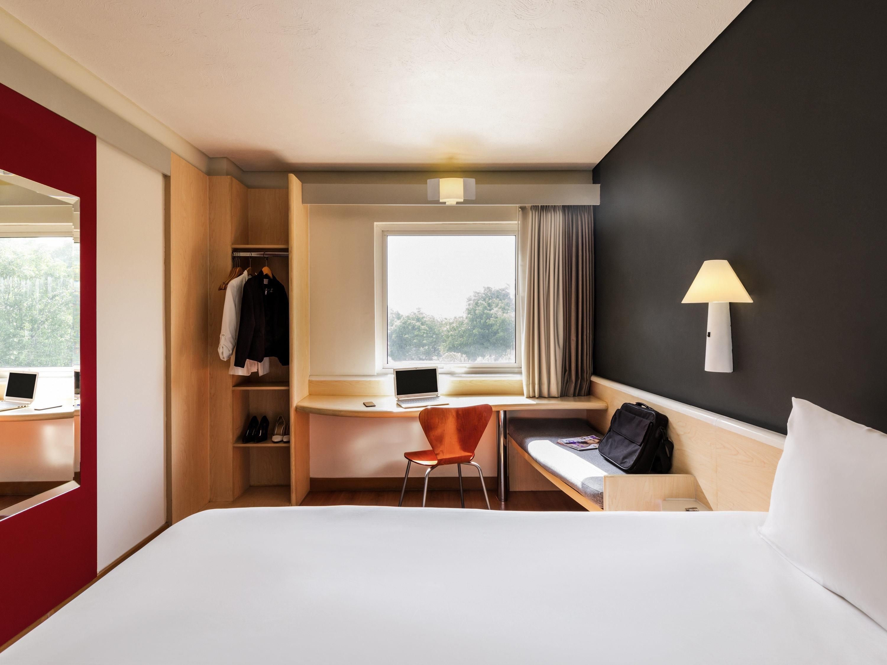 A hotel room with a bed, a desk with a laptop, a chair, a wardrobe, and a window with curtains.