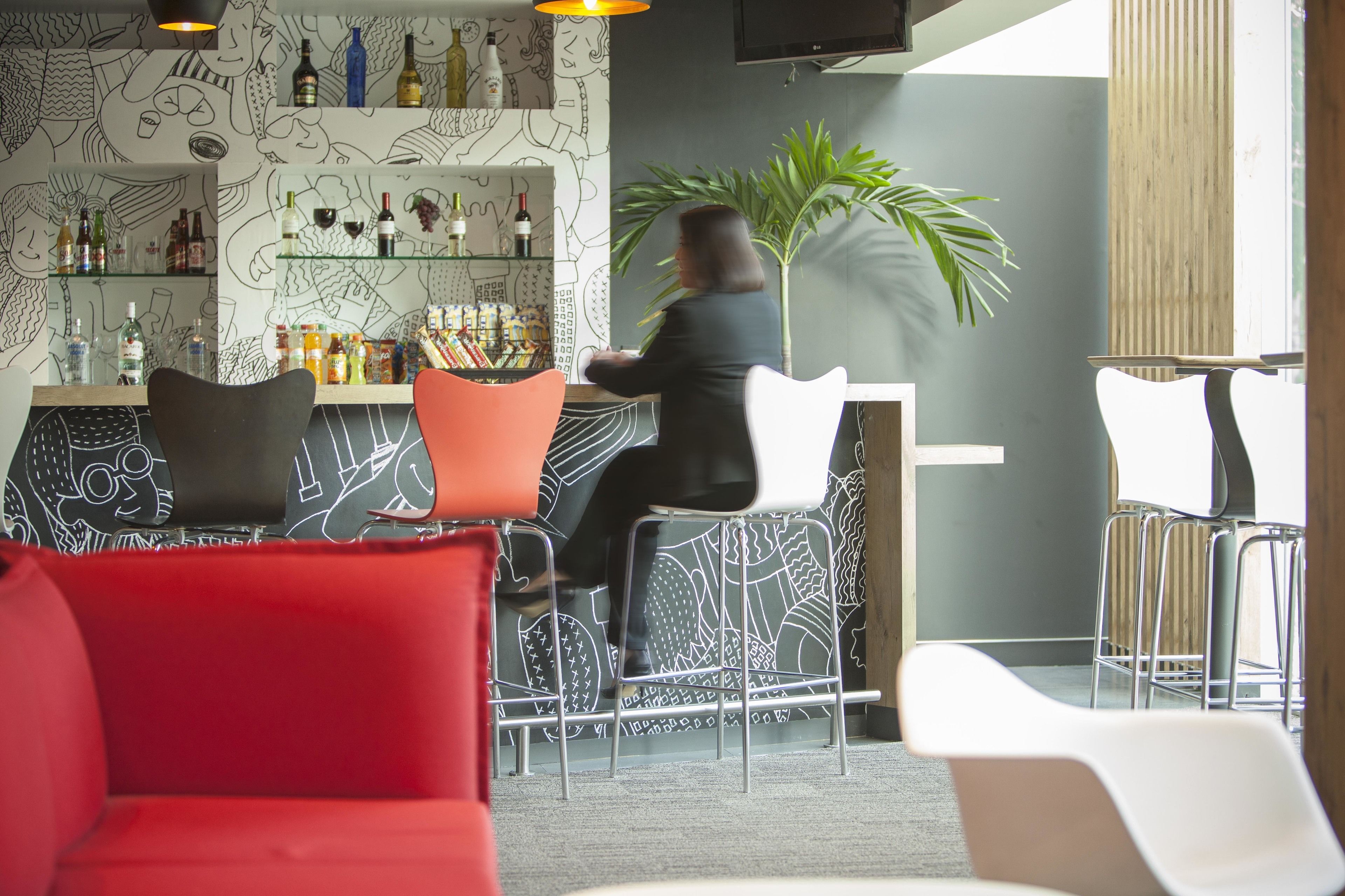 Un espace bar comprenant un comptoir avec un barman, des tabourets de bar, des étagères remplies de bouteilles et une grande plante.