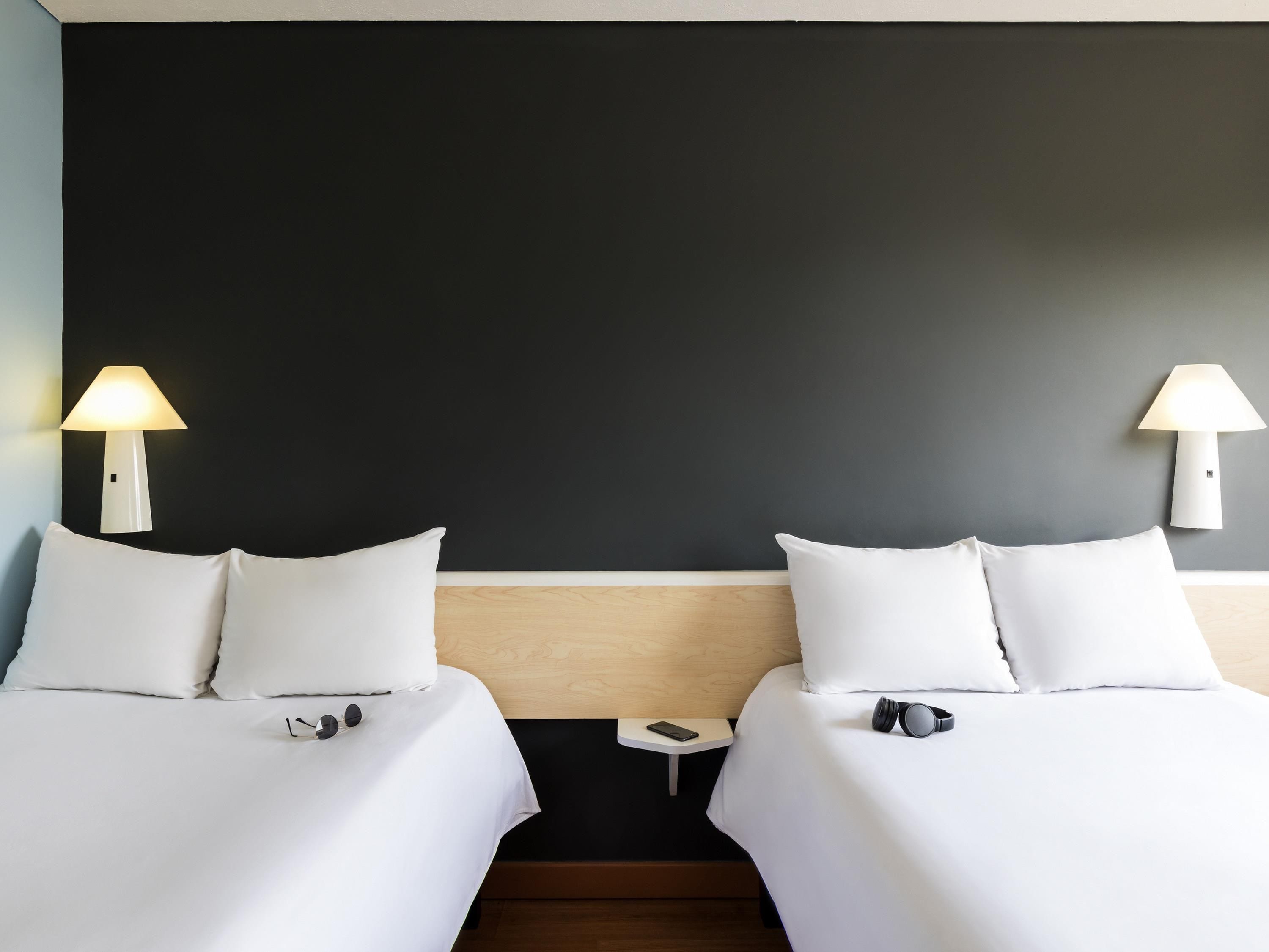 Une chambre d’hôtel avec deux lits, chacun doté de draps et d’oreillers blancs, une petite table de chevet avec une lampe et un smartphone posé sur la table de chevet.
