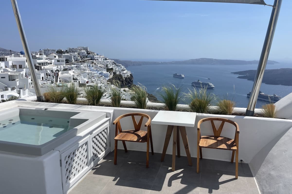 Una terraza con piscina, dos sillas de madera y una mesa, con vistas a un pueblo costero de edificios blancos y barcos en el agua.