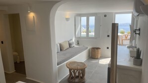 A modern, well-lit room with a built-in bench, a small round wooden stool, and a view of the outdoors through large windows. - Nomikos Villas (Santorini)