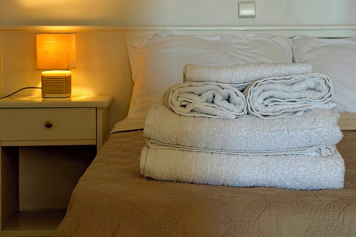 Una cama bien tendida con toallas dobladas, una lámpara de noche y una mesita de noche con cajón.
