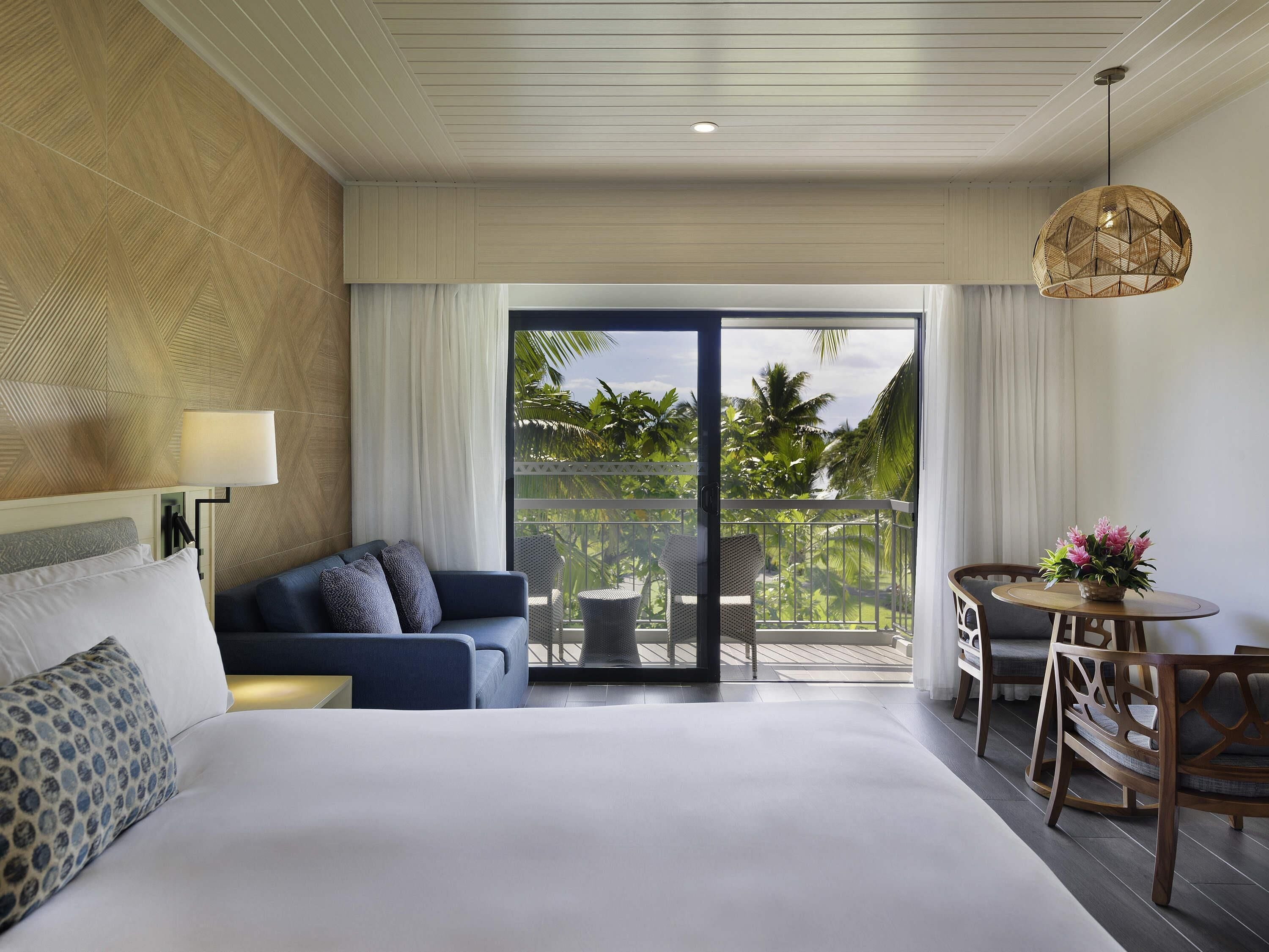 A hotel room with a bed, a sofa, a dining table, and a balcony with a view of palm trees.