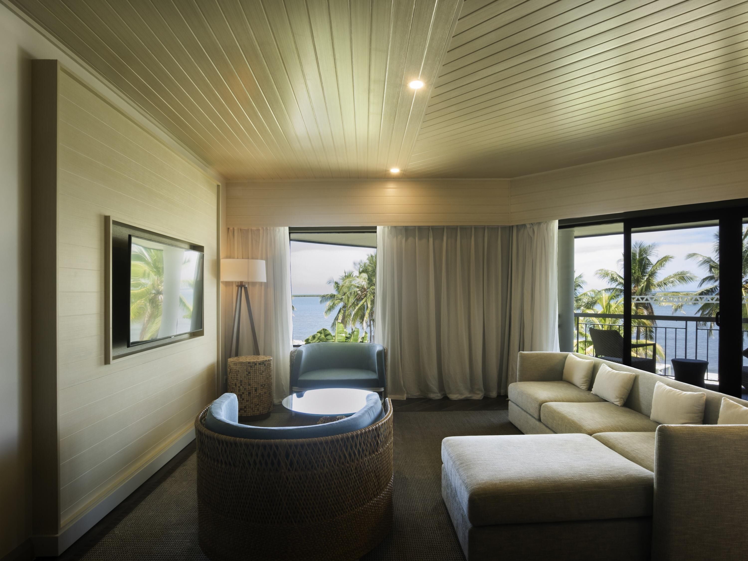 A hotel room with a sofa, a chair, a round table, a lamp, a TV, and a view of the ocean.