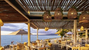 An outdoor dining area with tables and chairs, a pergola, hanging wicker lamps, and a view of the sea.