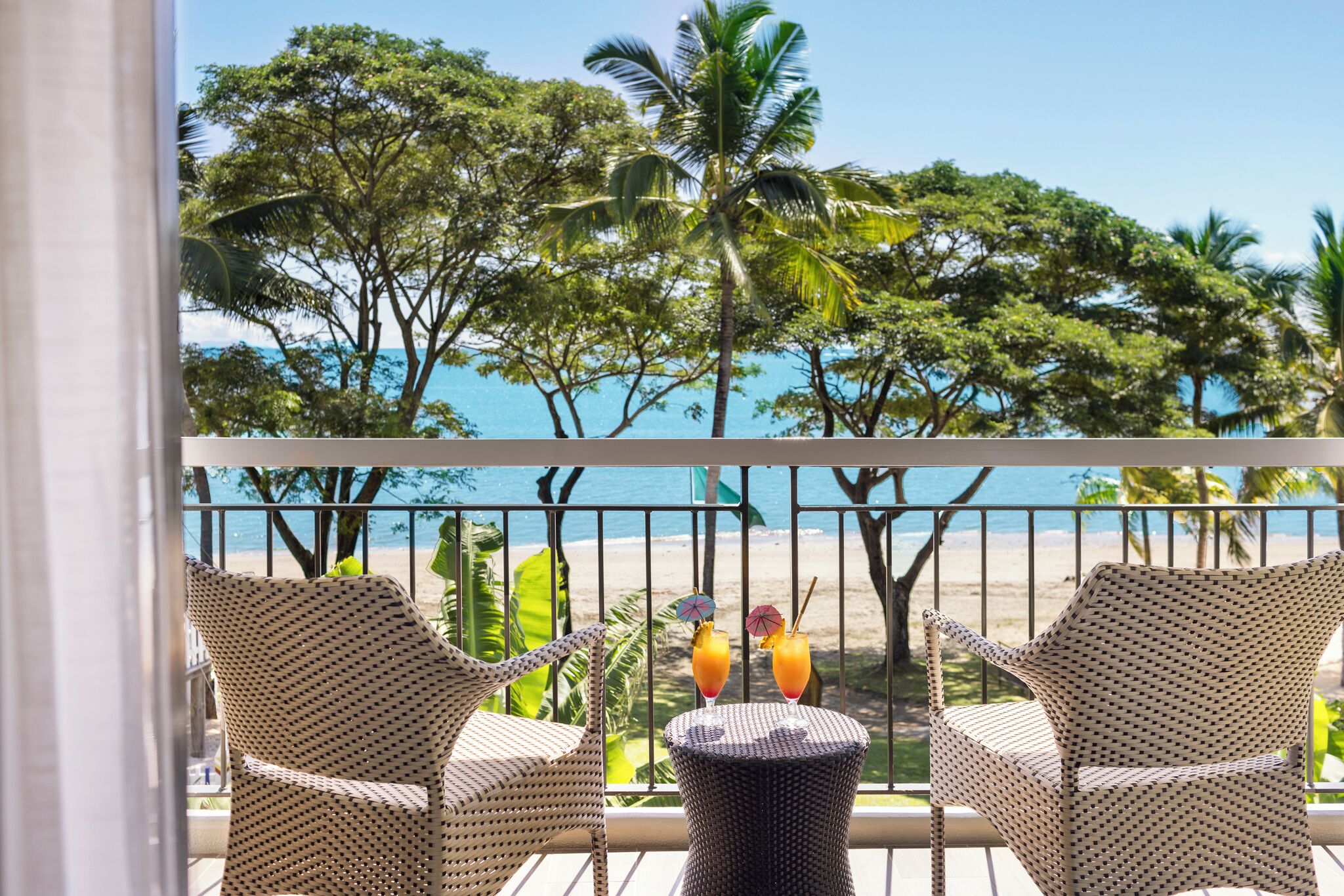 A balcony with wicker chairs and a small round table, overlooking a beach with palm trees and the ocean.