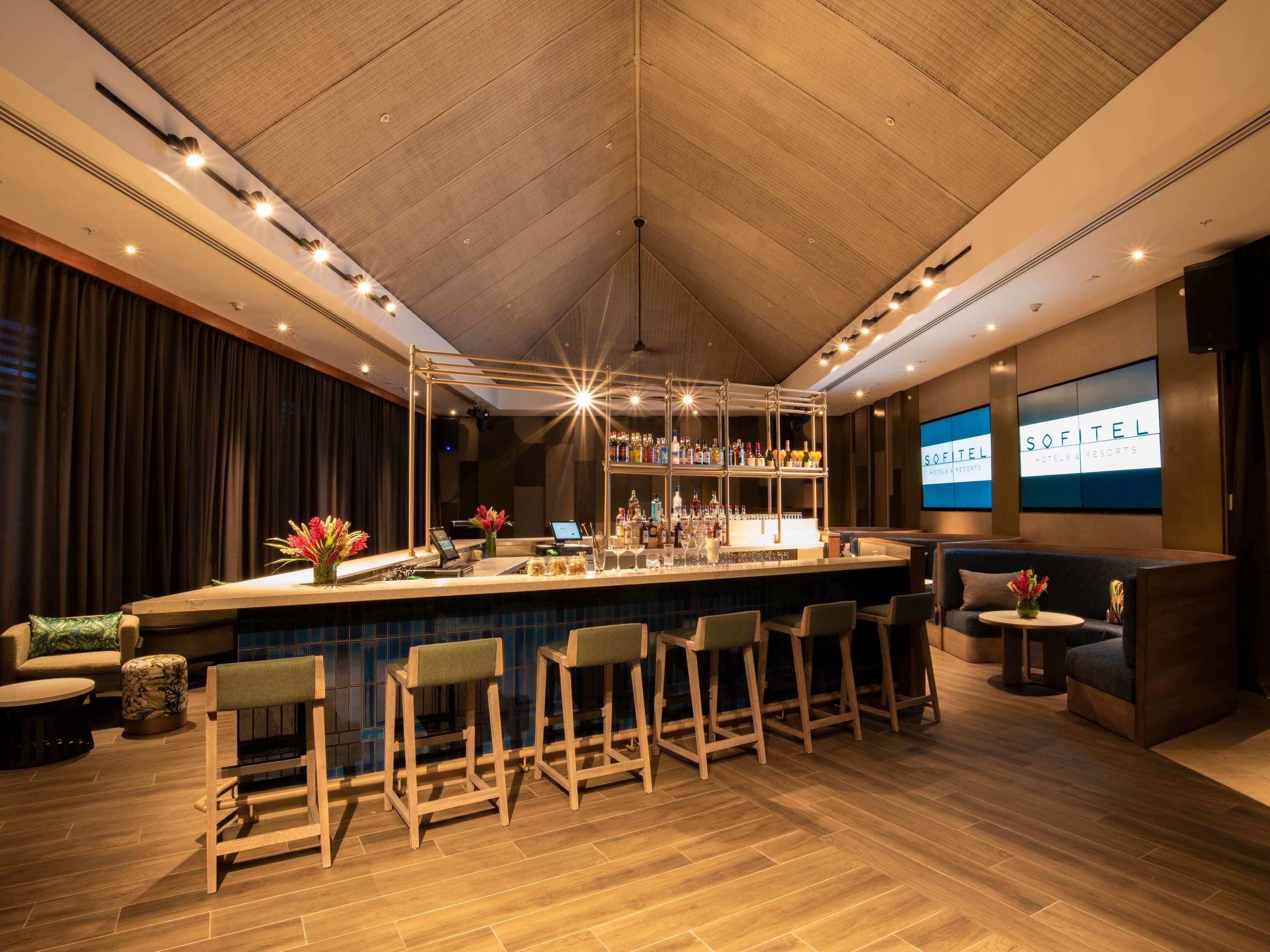 A bar area with a wooden ceiling, bar stools, a counter with a wine cooler, and a screen displaying 'SOFITEL HOTELS & RESORTS'.
