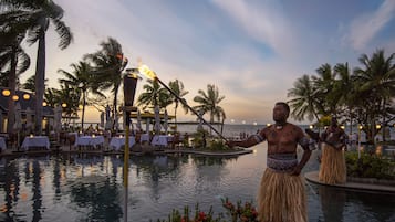A poolside area with tables and chairs, palm trees, and a view of the ocean.
