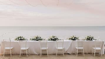 Mobiliario de comedor frente a la playa con manteles blancos, sillas y centros de mesa florales.