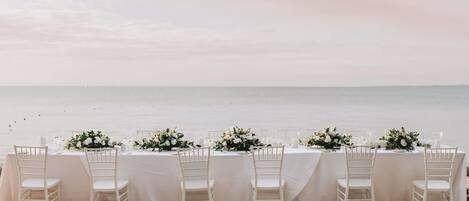 Mobiliario de comedor frente a la playa con manteles blancos, sillas y centros de mesa florales.
