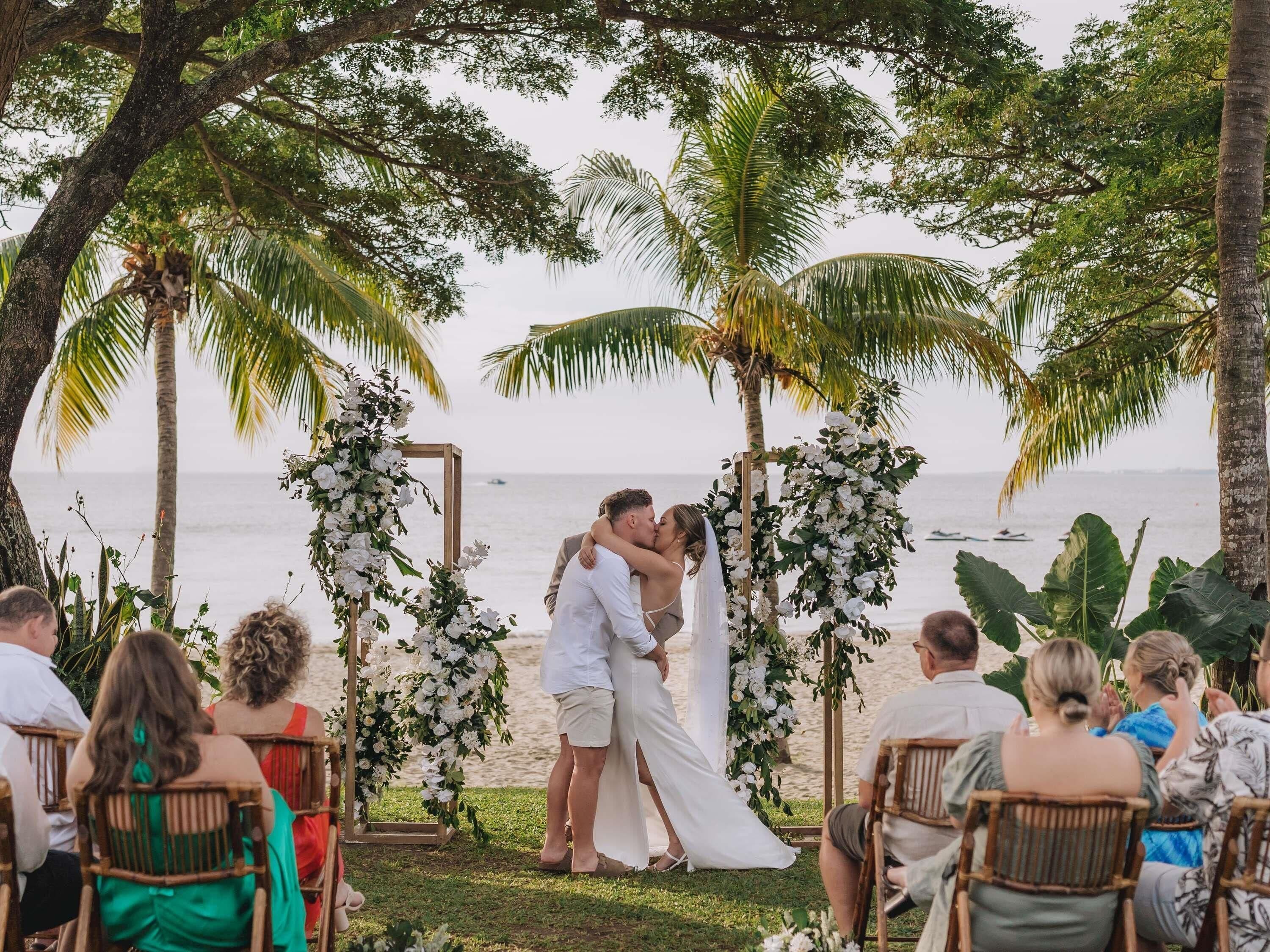 Una ceremonia de boda con una pareja besándose bajo un arco floral en la playa.