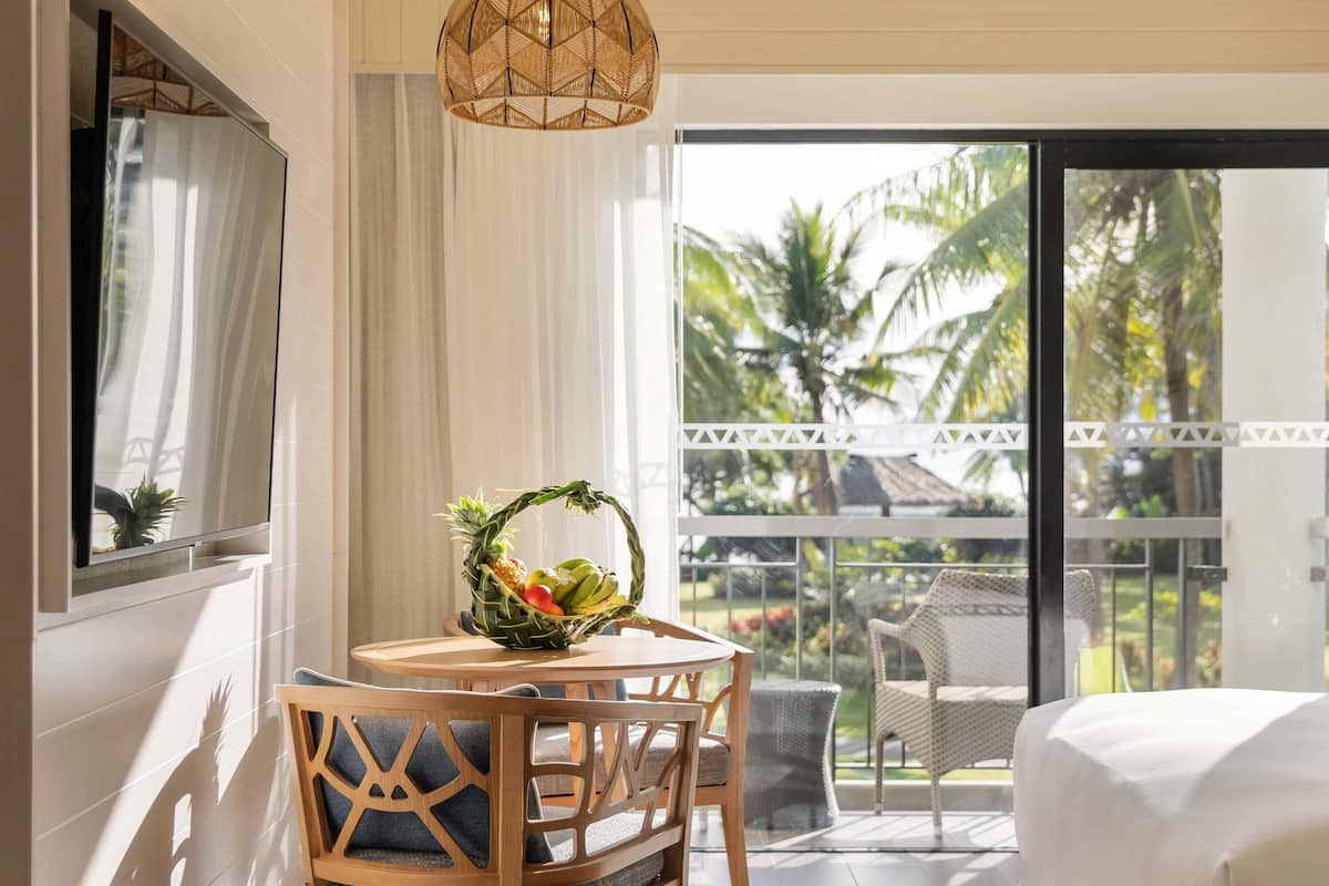 A hotel room with a dining table, chairs, a fruit basket, a bed, and a balcony with a view of palm trees.