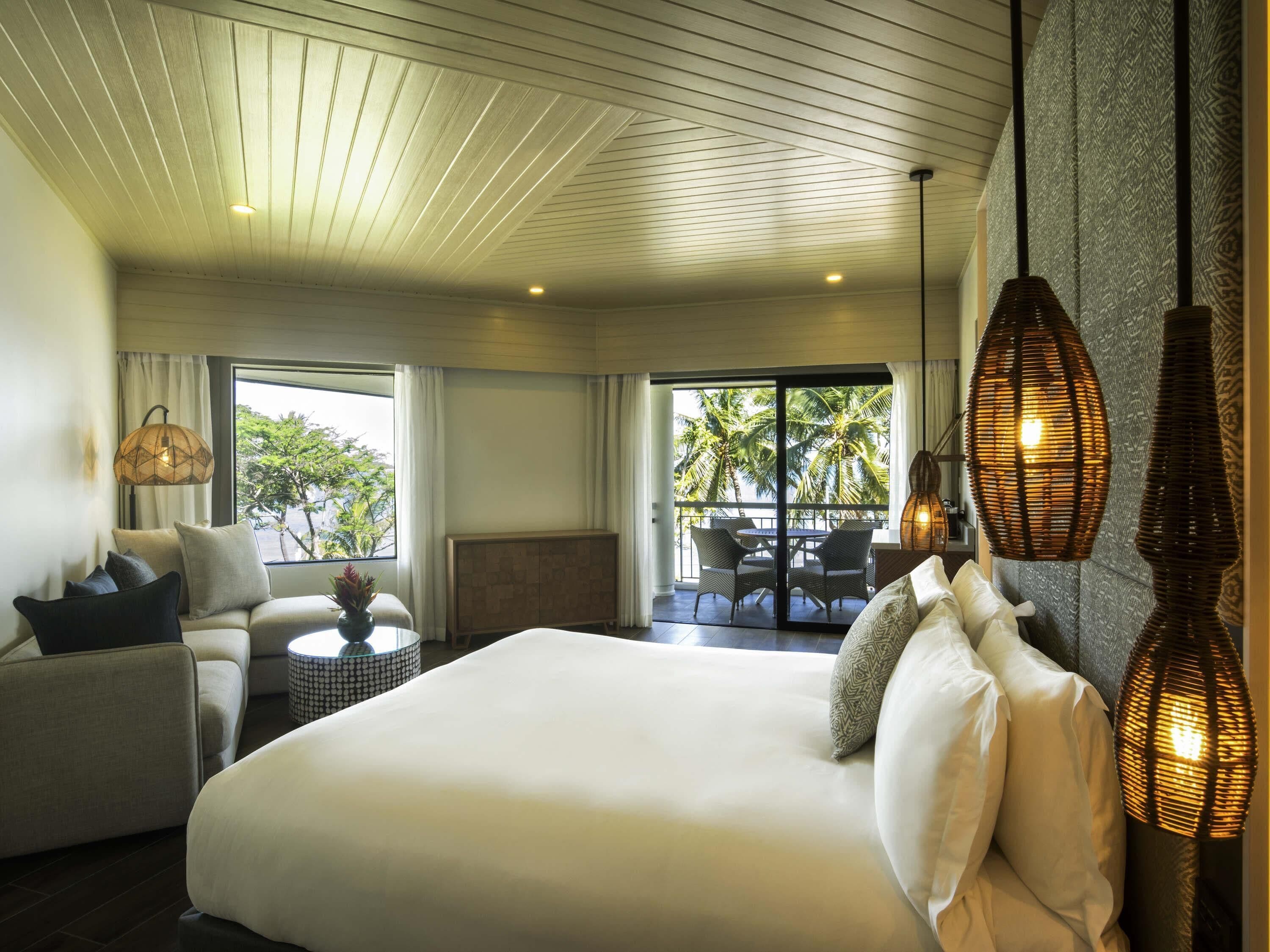 A hotel room with a large bed, a sofa, a dining table, and a balcony with a view of palm trees.