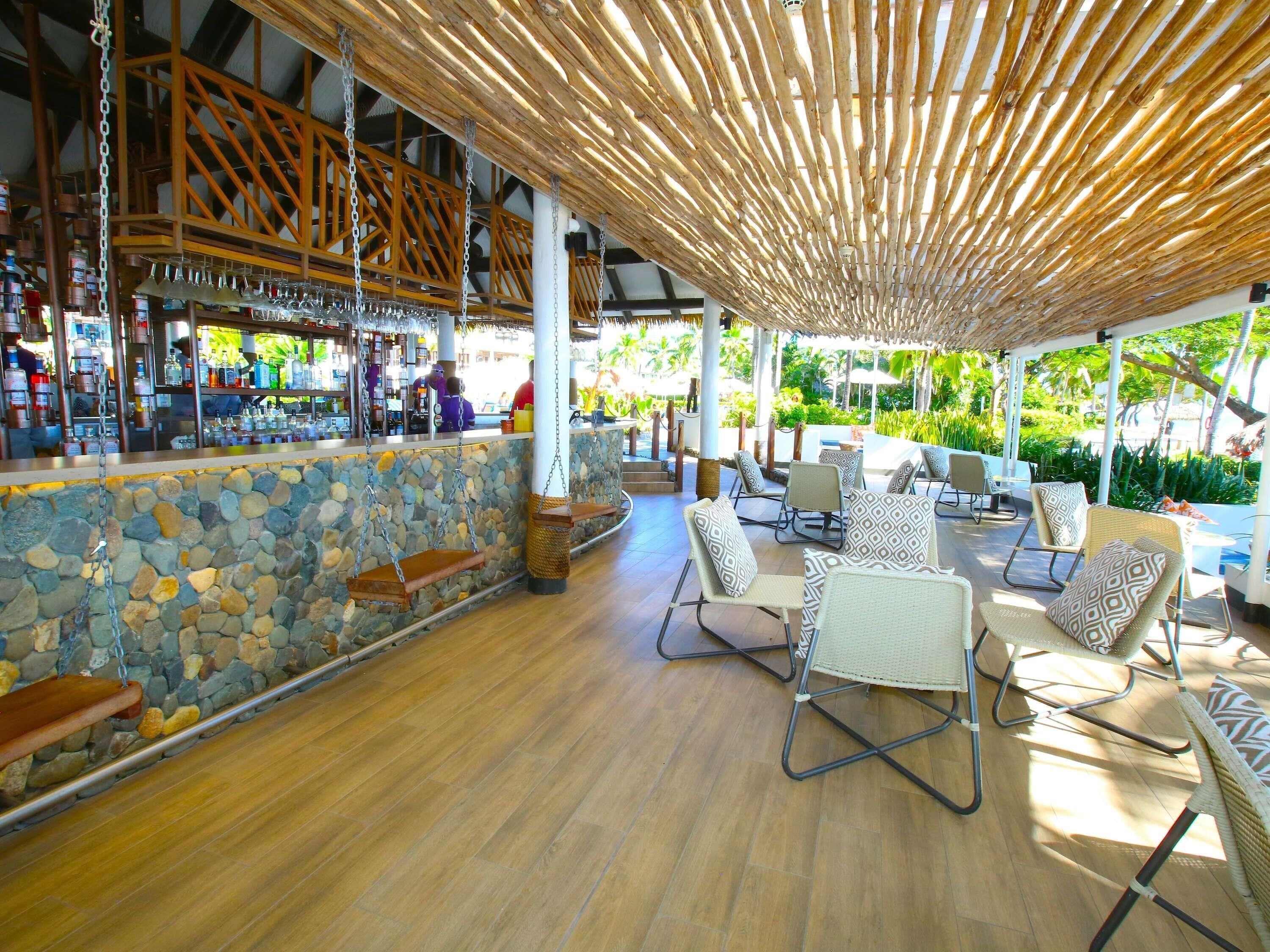 Zona de bar con pared de piedra, techo de madera y asientos exteriores con cojines estampados.