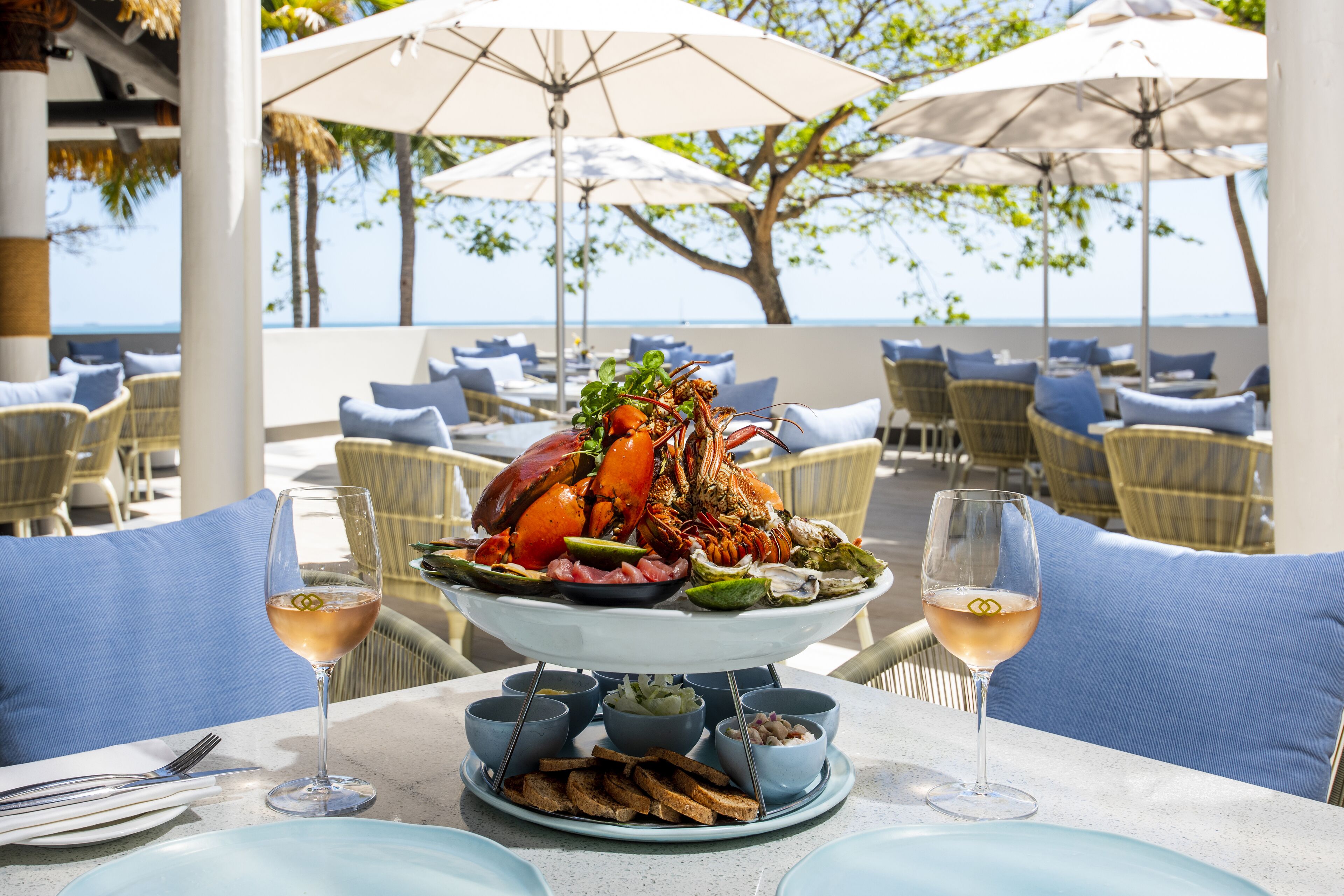 A seafood platter on a table with wine glasses, surrounded by wicker chairs and umbrellas, overlooking a beach.
