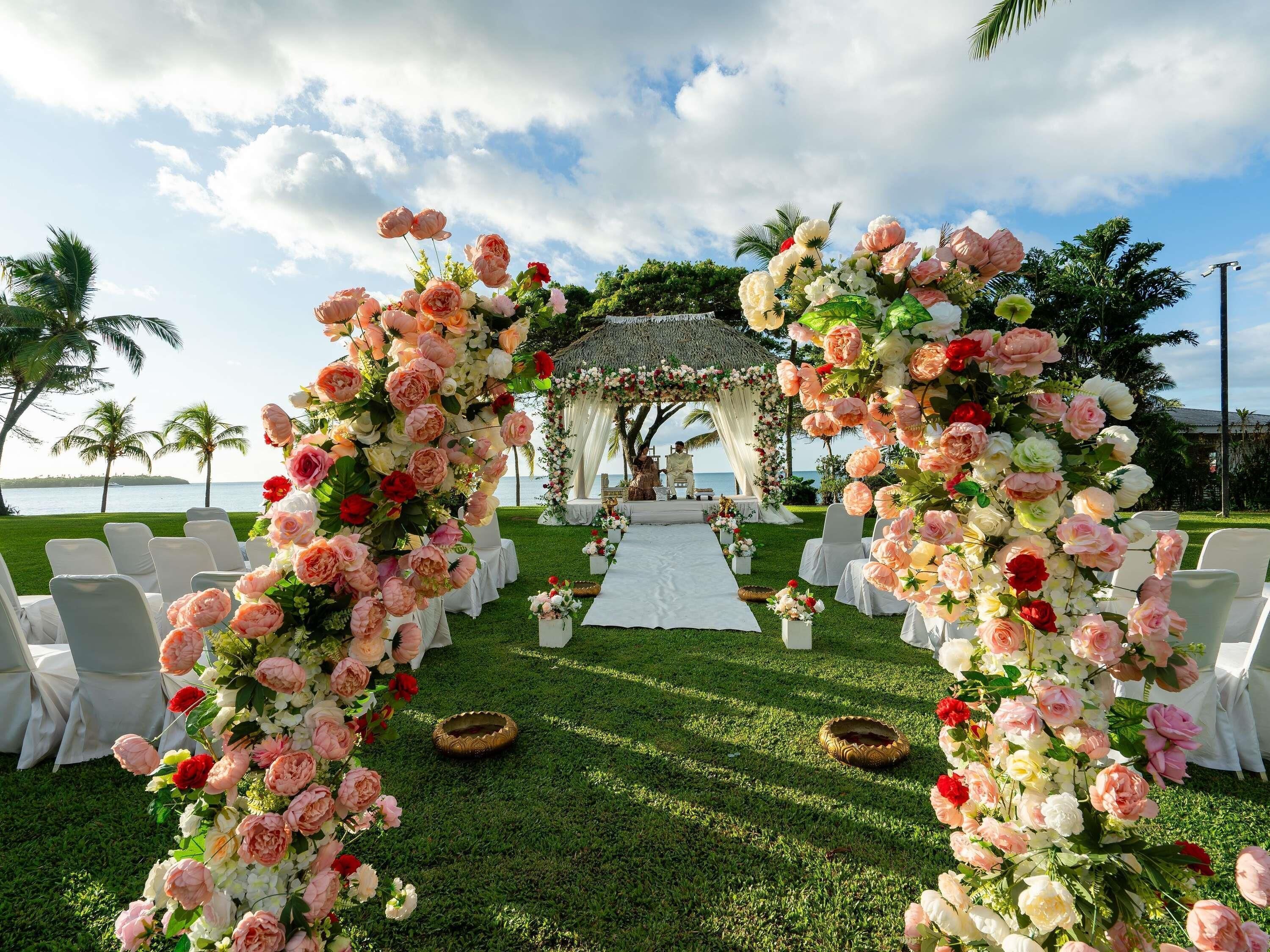 Espaço para casamento ao ar livre com arco de flores, cadeiras brancas e mesa coberta por toalha branca.