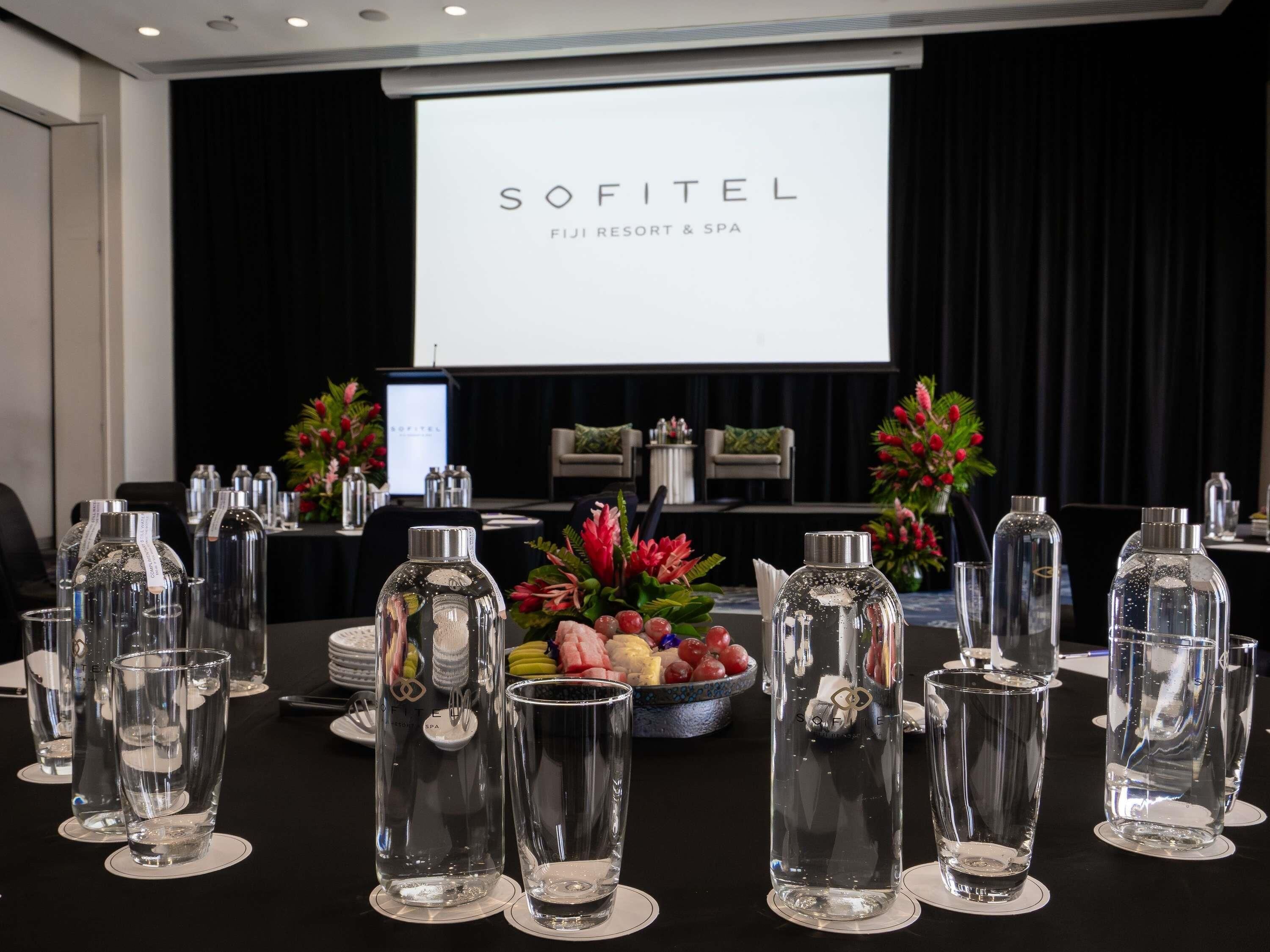 A conference room with a projection screen displaying 'SOFITEL FIJI RESORT & SPA', a table set with clear glasses and a fruit bowl, and a black curtain backdrop.
