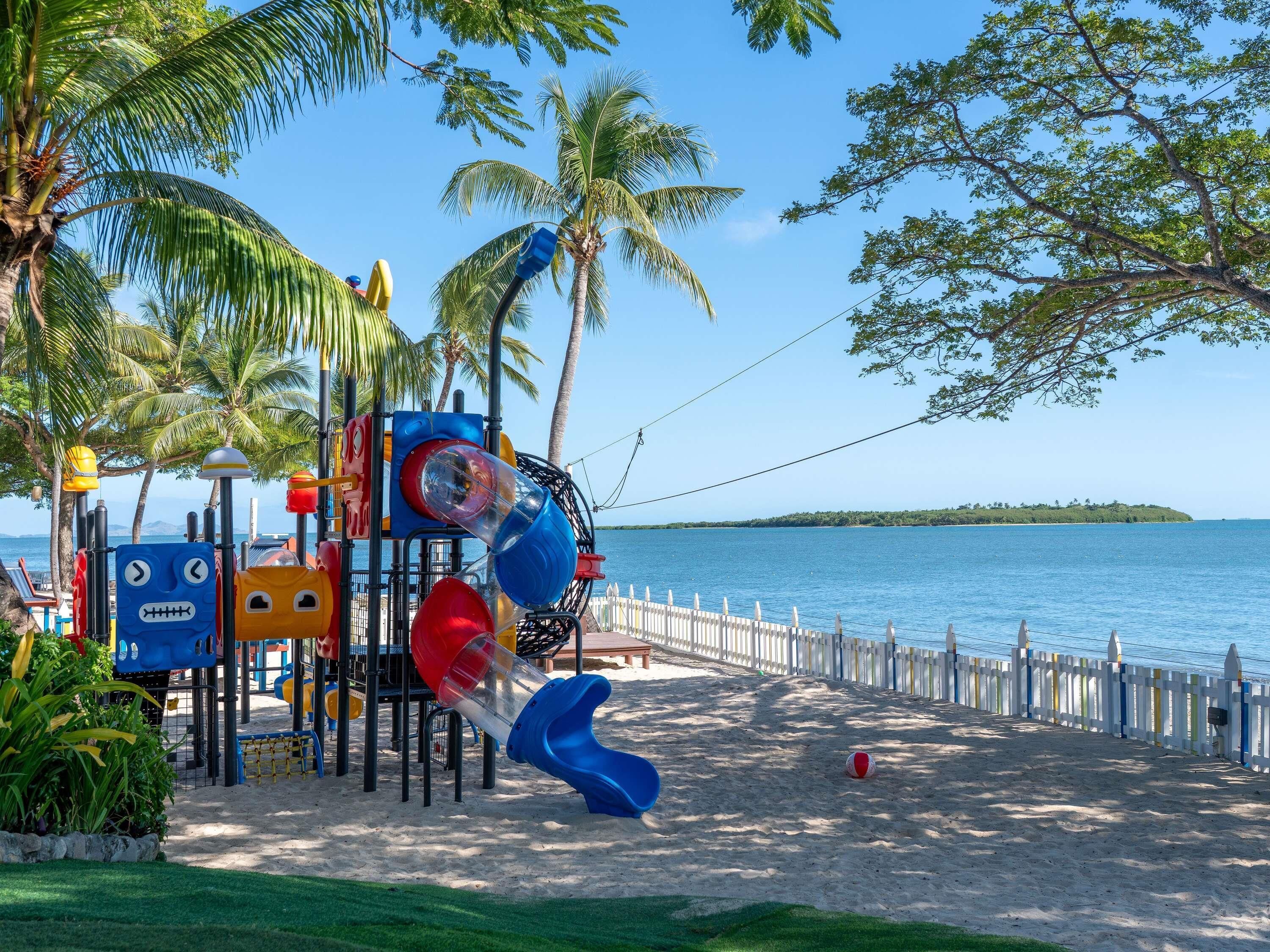 Un parque infantil con toboganes y estructuras para escalar cerca de una playa con vallas y palmeras.