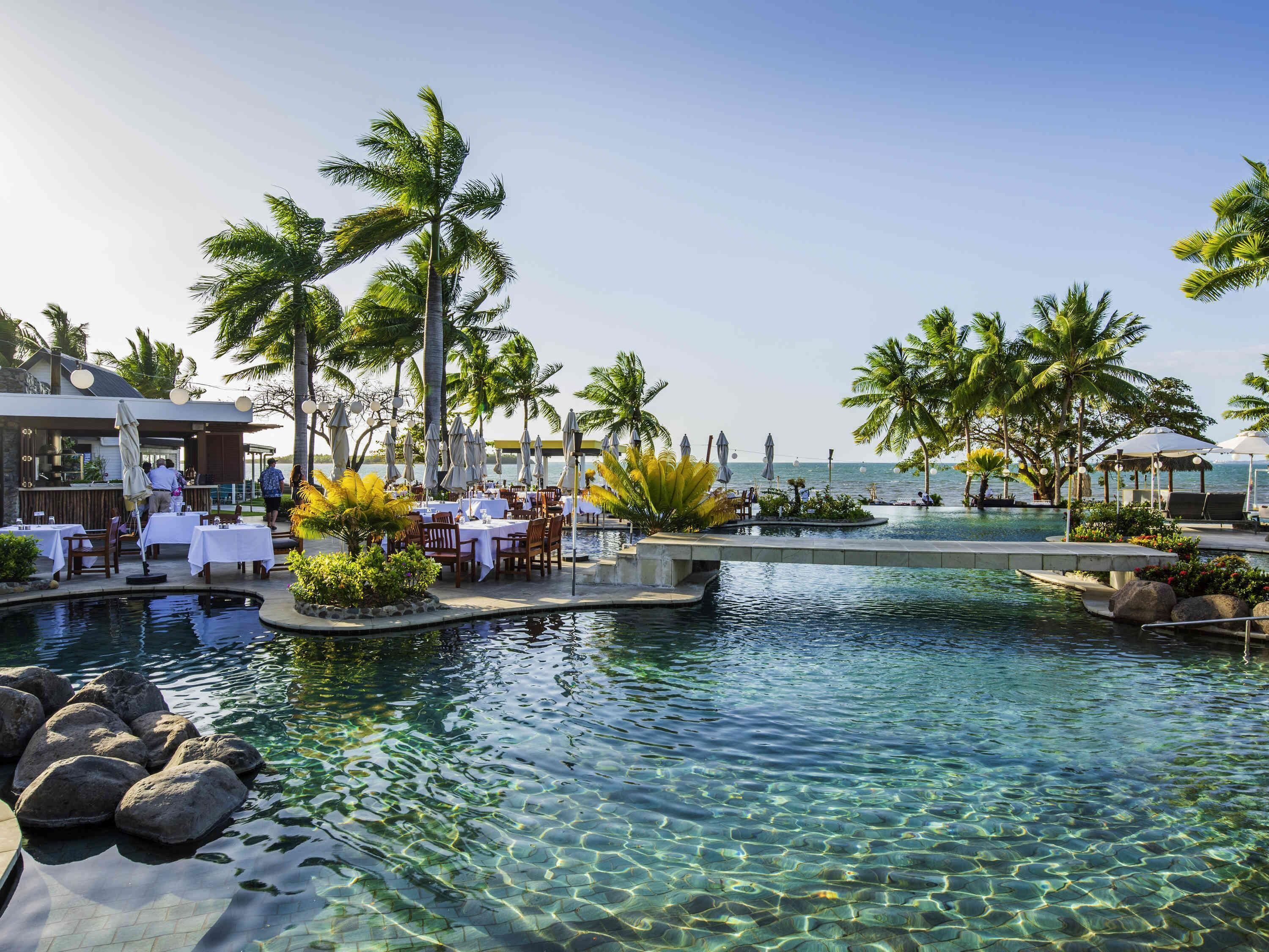 A resort pool area with clear water, palm trees, and outdoor dining tables.