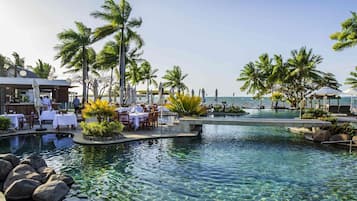 A resort pool area with clear water, palm trees, and outdoor dining tables.