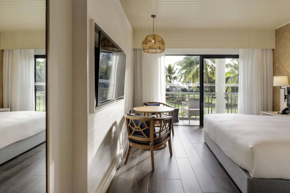 Une chambre d’hôtel avec un lit, une table à manger avec des chaises, une fenêtre avec des rideaux et une vue sur des palmiers.