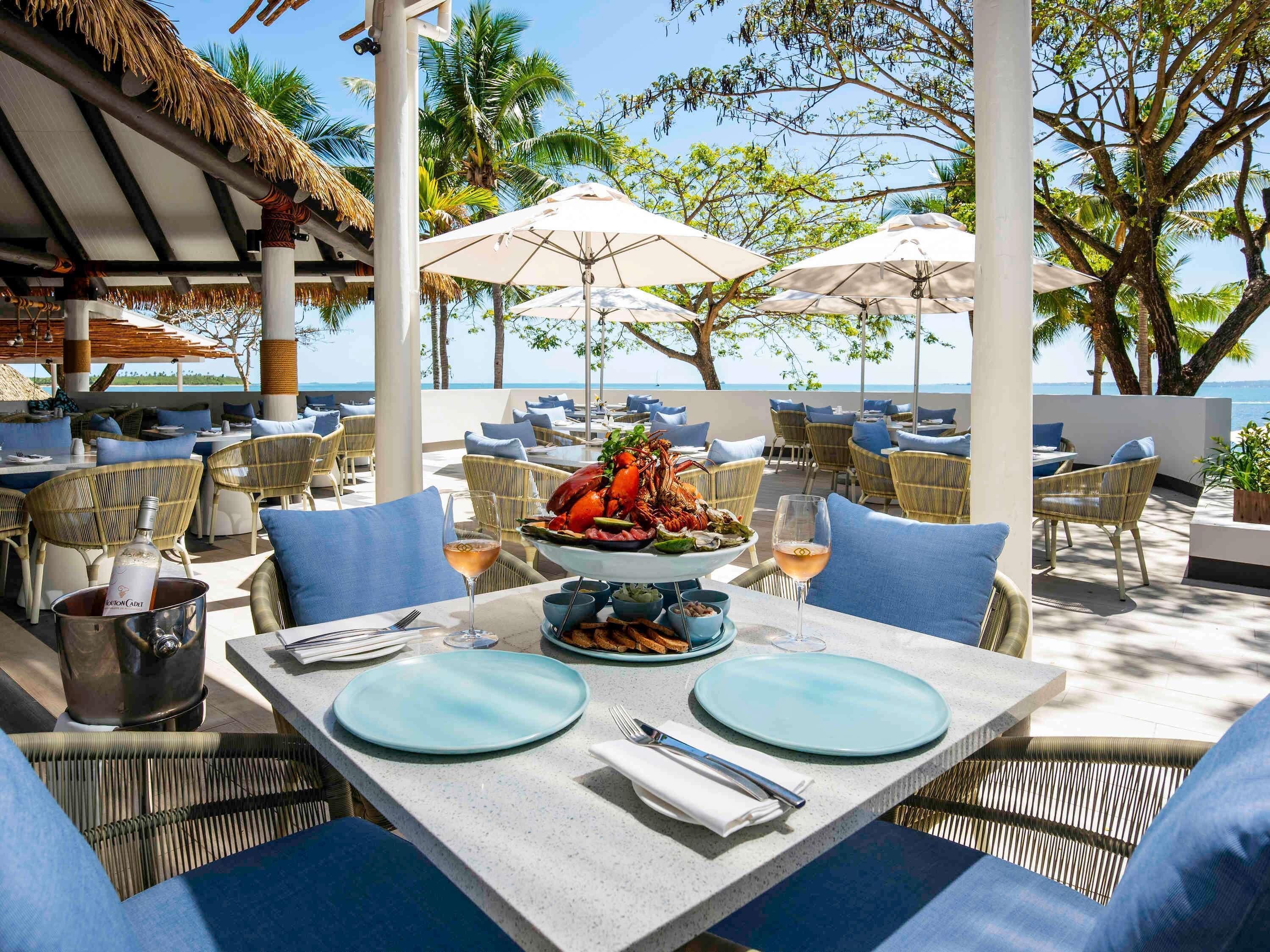 Un restaurant en bord de mer avec une terrasse, un plat de homard, des verres à vin et une vue sur l’océan.