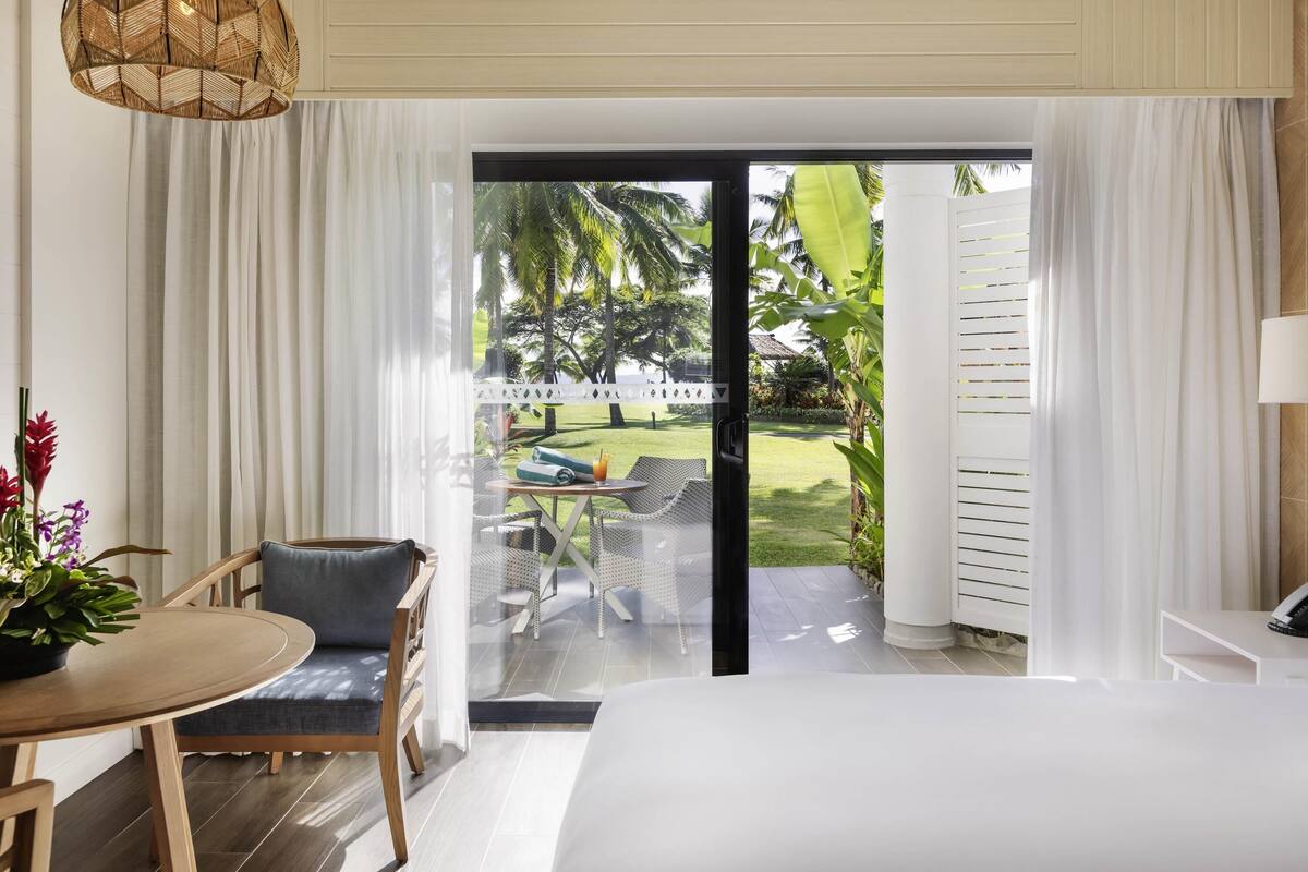 Une chambre d’hôtel avec un lit, une table à manger avec des chaises, un luminaire suspendu en osier et une vue sur un jardin avec une piscine et des palmiers.