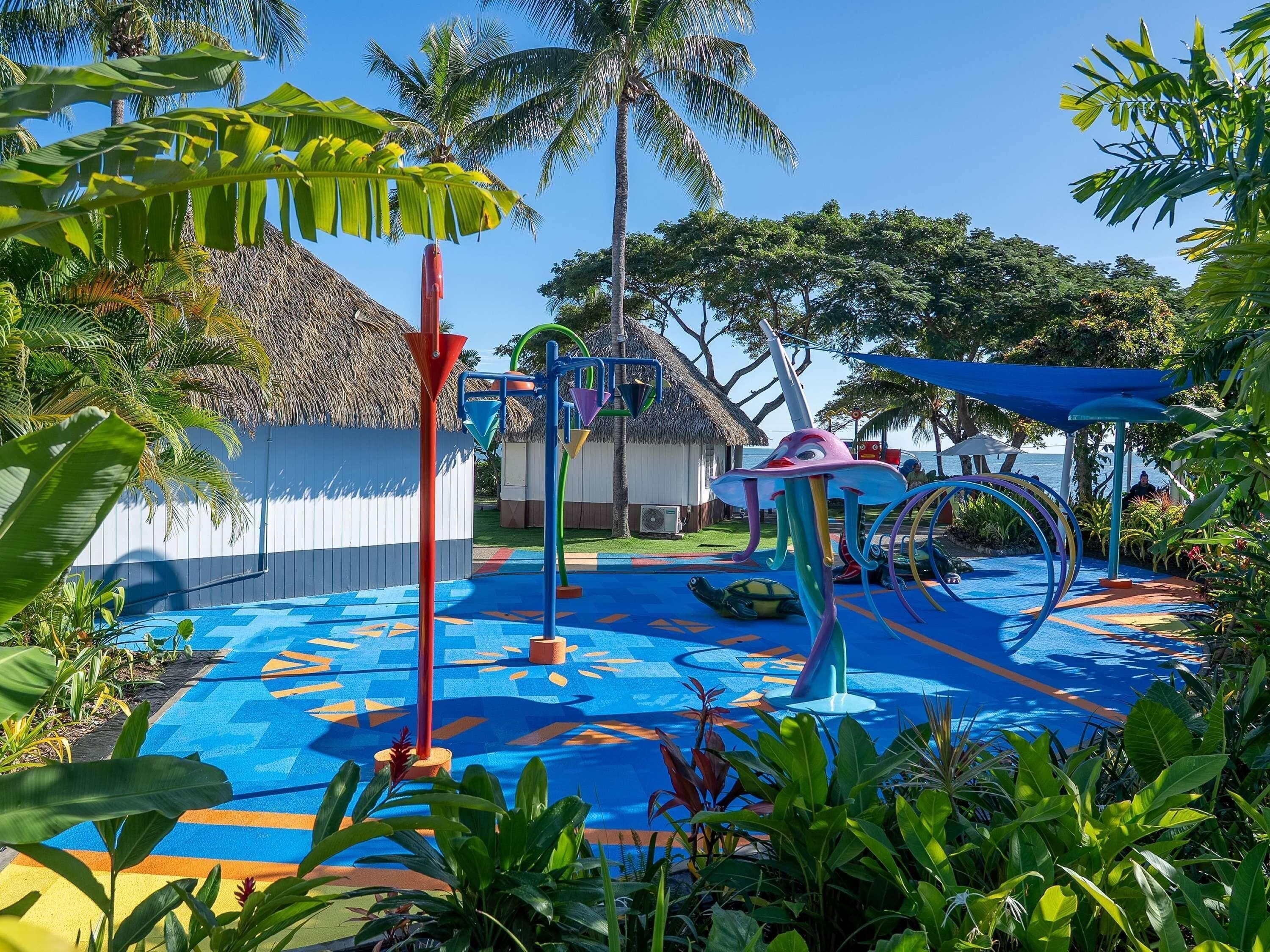 A tropical playground with colorful play structures, a thatched-roof hut, and lush greenery.